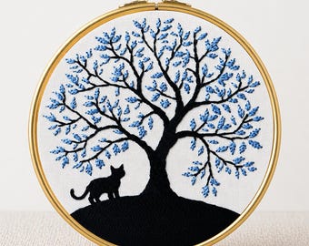 May include: An embroidery hoop featuring a black tree with blue leaves and a black cat silhouette. The design is set against a white background and framed by a gold hoop. The text "PDF EMBROIDERY PATTERN" is at the bottom.