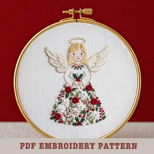 May include: Embroidered angel hoop art with a gold frame. The angel has blonde hair, a halo, and white wings. Her dress is adorned with red roses, white flowers, and green leaves. The text "PDF EMBROIDERY PATTERN" is at the bottom.