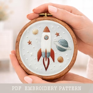 Op de afbeelding: Geborduurde ruimte-thema met een witte en rode raket, planeten en sterren op een lichtblauwe achtergrond, in een houten hoepel. De tekst "PDF EMBROIDERY PATTERN" staat onderaan.