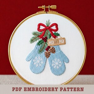May include: Embroidered Christmas design featuring light blue mittens with snowflake details, a red bow, and a sprig of greenery. A small sign reads "HOHOHO". The design is set in a gold embroidery hoop. PDF Embroidery Pattern.