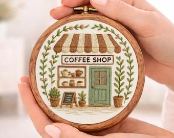 Puede incluir: Un bastidor de bordado redondo de madera con una detallada escena de cafetería. El diseño incluye un toldo a rayas, una puerta verde y el texto "COFFEE SHOP". El bordado está rodeado de hojas verdes y pequeñas flores rosas.