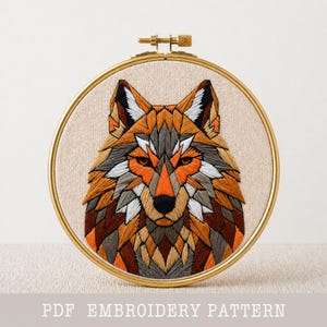Peut inclure: Broderie d'une tête de loup dans un cercle doré. Le loup est représenté avec des formes géométriques dans des tons orange, marron, gris et blanc. Le texte "PDF EMBROIDERY PATTERN" est au bas de l'image. La broderie est sur un tissu beige.