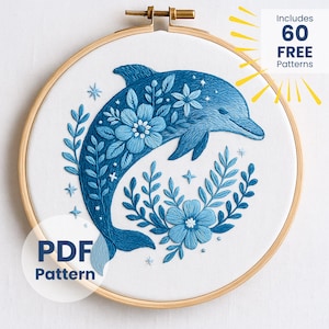 Puede incluir: Diseño de delfín bordado en un aro de madera. El delfín es azul, rodeado de flores y follaje azules. El texto "Includes 60 FREE Patterns" está en la esquina superior derecha. Hay un patrón PDF disponible.