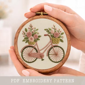 Pode incluir: Um bastidor de bordado redondo com um design de bicicleta rosa, completo com cestos de rosas cor-de-rosa e folhas verdes. O bordado está sobre tecido creme, emoldurado num aro de madeira. O texto "PDF EMBROIDERY PATTERN" está na parte inferior.