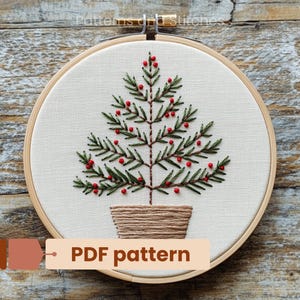 Könnte beinhalten: Stickmuster eines Weihnachtsbaums in einem Reifen. Der Baum ist grün mit roten Beeren und in einem braunen Topf eingetopft. Der Text "PDF pattern" befindet sich unter dem Reifen.