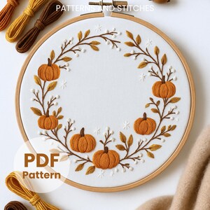 May include: An embroidery hoop containing white fabric with a circular wreath design featuring orange pumpkins, brown stems, and small white berries. The text PATTERNS AND STITCHES and PDF Pattern are visible. Several skeins of embroidery thread are nearby.
