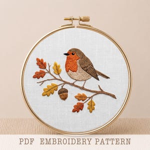 May include: An embroidery hoop containing white fabric with a hand-stitched robin perched on a brown branch. The branch features orange and yellow oak leaves and a single brown acorn. The text PDF EMBROIDERY PATTERN is printed below the hoop.