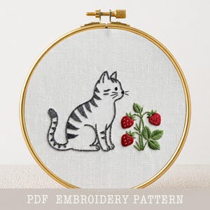 May include: Embroidered cat and strawberry design in a gold hoop. The cat is gray and white with black stripes. The strawberries are red with green leaves. The text "PDF EMBROIDERY PATTERN" is at the bottom.