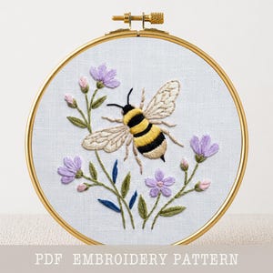 May include: A hand-stitched embroidery hoop featuring a bee with yellow and black stripes, surrounded by purple flowers and green leaves. The embroidery is set against a white fabric background, framed by a gold hoop. The text "PDF EMBROIDERY PATTERN" is at the bottom.