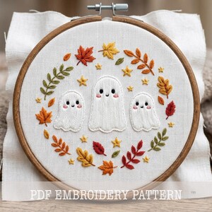 May include: A circular wooden embroidery hoop containing white fabric with a hand-stitched design of three white ghosts surrounded by a wreath of colorful autumn leaves and small yellow stars. The text PDF EMBROIDERY PATTERN is visible.