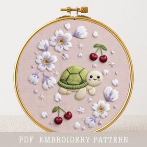 May include: A hand-stitched embroidery hoop featuring a cute turtle with a green shell, surrounded by white and purple flowers, and red cherries. The design is set against a pink fabric background, framed by a gold hoop. Text at the bottom reads "PDF EMBROIDERY PATTERN".