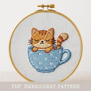 Pode incluir: Um bastidor de bordado com um gato laranja bordado numa chávena azul clara com bolinhas brancas. O gato tem os olhos fechados e uma cauda listrada. O texto "PDF EMBROIDERY PATTERN" está na parte inferior.