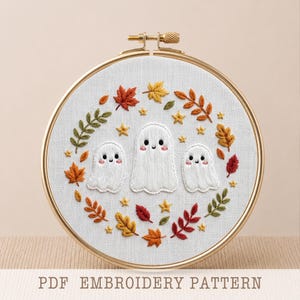 May include: A circular wooden embroidery hoop containing white fabric with a hand-stitched design of three white ghosts surrounded by a wreath of colorful autumn leaves and small yellow stars. The text PDF EMBROIDERY PATTERN is visible below.