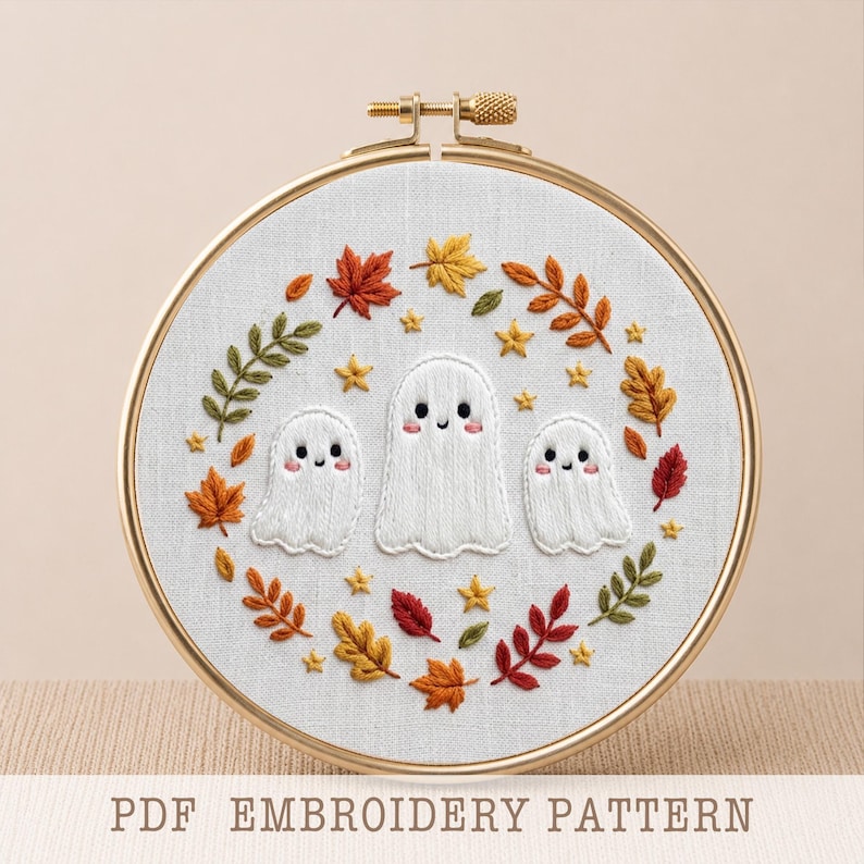 May include: A circular wooden embroidery hoop containing white fabric with a hand-stitched design of three white ghosts surrounded by a wreath of colorful autumn leaves and small yellow stars. The text PDF EMBROIDERY PATTERN is visible below.