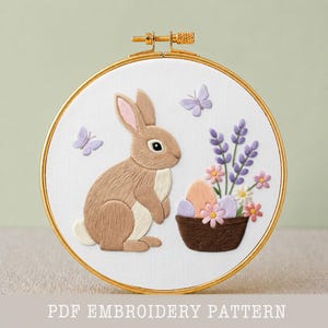 May include: Embroidered Easter-themed hoop art featuring a brown bunny, butterflies, and a basket of eggs and flowers. The design is set against a white background within a gold-toned hoop. The text "PDF EMBROIDERY PATTERN" is at the bottom.