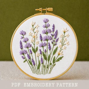 May include: A round embroidery hoop with a white fabric featuring a floral design. The design includes purple lavender, white daisies, and green leaves. The text "PDF EMBROIDERY PATTERN" is at the bottom.