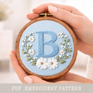 Può includere: Ricamo su telaio con la lettera "B" azzurra, circondata da margherite bianche e foglie verdi su tessuto azzurro chiaro. Il design è racchiuso in un telaio di legno. Il testo "PDF EMBROIDERY PATTERN" è in basso.