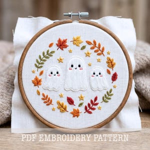 May include: An embroidery hoop containing white fabric with a hand-stitched design of three white ghosts with pink cheeks surrounded by a wreath of colorful autumn leaves and small yellow stars. The text PDF EMBROIDERY PATTERN is visible.
