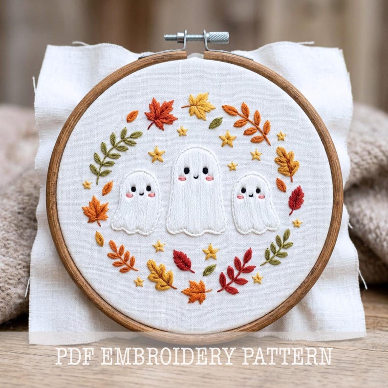May include: An embroidery hoop containing white fabric with a hand-stitched design of three white ghosts with pink cheeks surrounded by a wreath of colorful autumn leaves and small yellow stars. The text PDF EMBROIDERY PATTERN is visible.
