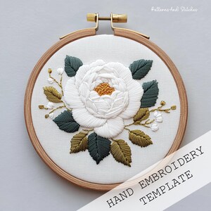 Peut inclure: Modèle de broderie à la main représentant une fleur blanche avec un centre doré, entourée de feuilles vertes et dorées, et de petites fleurs blanches. Le motif est placé dans un cercle à broder en bois. Le texte "HAND EMBROIDERY TEMPLATE" est visible.
