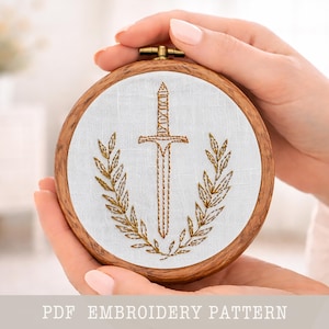 May include: A round wooden embroidery hoop with a white fabric featuring a gold embroidered design of a sword framed by laurel leaves. The text "PDF EMBROIDERY PATTERN" is at the bottom.