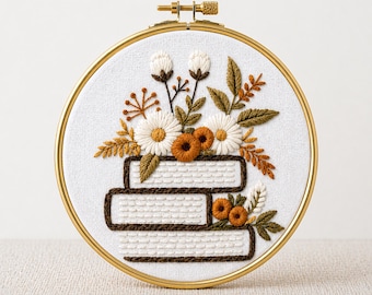 May include: A round embroidery hoop with a gold-colored frame. The white fabric inside features an embroidered design of stacked books with flowers and foliage in shades of white, orange, and brown. The text "PDF EMBROIDERY PATTERN" is at the bottom.