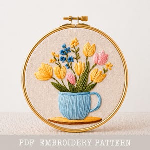 Peut inclure: Broderie florale dans un cercle doré. L'œuvre présente des fleurs jaunes, roses et bleues disposées dans une tasse bleue sur un dessous de plat en bois. Le texte "PDF EMBROIDERY PATTERN" est en bas.