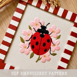 Peut inclure: Broderie d'une coccinelle rouge à pois noirs, entourée de fleurs roses et de feuilles vertes. L'œuvre est encadrée dans un cadre en bois à rayures rouges et blanches. Le texte "PDF EMBROIDERY PATTERN" est en bas.