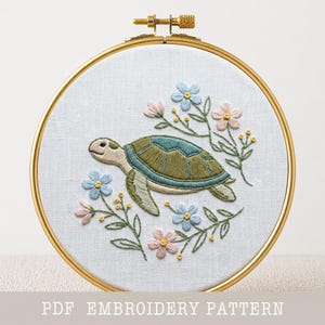 Könnte beinhalten: Gesticktes Schildkröten-Design in einem goldenen Reifen. Die Schildkröte hat einen grünen und blauen Panzer, umgeben von rosa und blauen Blumen und grünen Blättern. Der Text "PDF EMBROIDERY PATTERN" steht unten.