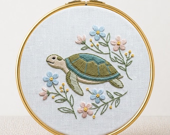 May include: Embroidered turtle design in a gold hoop. The turtle has a green and blue shell, surrounded by pink and blue flowers and green leaves. The text "PDF EMBROIDERY PATTERN" is at the bottom.