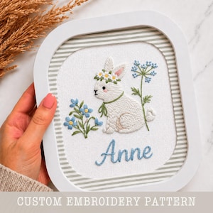 Puede incluir: Obra bordada con un conejito blanco con una corona de flores y un lazo verde, rodeado de flores azules. El nombre "Anne" está bordado en azul. La obra está enmarcada en un marco blanco cuadrado con un fondo a rayas. El texto "CUSTOM EMBROIDERY PATTERN" está en la parte inferior.