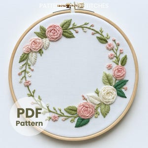 Könnte beinhalten: Ein floraler Stickrahmen mit einem Kranz aus rosa und weißen Rosen, grünen Blättern und kleinen rosa Knospen. Die Stickerei befindet sich auf weißem Stoff in einem Holzreifen. Der Text "PDF Pattern" ist sichtbar.