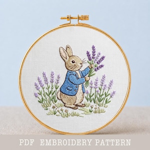 Op de afbeelding: Een borduurring met een Peter Konijn-ontwerp. De geborduurde afbeelding toont een bruin konijn met een blauw jasje, met lavendelbloemen. De achtergrond bevat lavendelplanten en gras. De tekst "PDF EMBROIDERY PATTERN" staat onderaan.