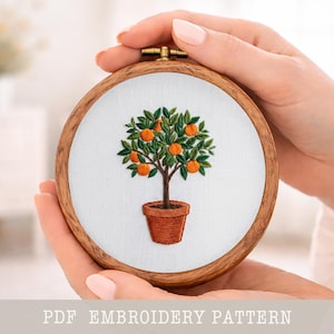May include: A round wooden embroidery hoop featuring a detailed tree with green leaves and orange fruits in a terracotta pot. The embroidery is set against a white background. The text "PDF EMBROIDERY PATTERN" is at the bottom.
