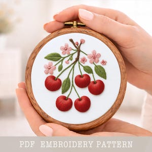 Puede incluir: Un bastidor bordado a mano con un diseño de cerezas. La obra presenta cinco cerezas rojas, hojas verdes y flores rosas sobre un fondo blanco, enmarcado en un aro de madera. El texto "PDF EMBROIDERY PATTERN" está en la parte inferior.