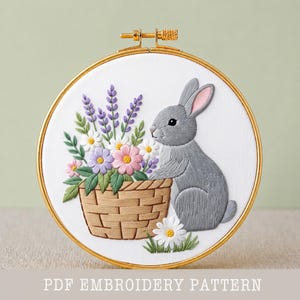 May include: An embroidered hoop art piece with a grey rabbit and a basket of flowers. The basket contains lavender, daisies, and pink and purple flowers. The text "PDF EMBROIDERY PATTERN" is at the bottom of the image.