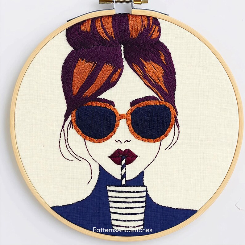 May include: Embroidery hoop art featuring a stylized woman with sunglasses and a drink. The woman's hair is purple and orange, and she wears orange-framed sunglasses with dark blue lenses. The drink cup is white with black stripes. The text "PatternsAndStitches" is at the bottom.