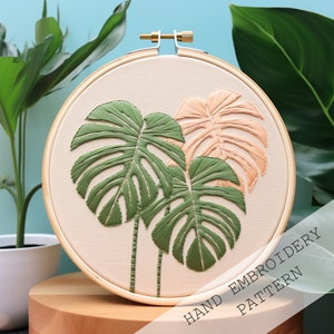 Pode incluir: Um padrão de bordado à mão com três folhas de monstera, duas verdes e uma pêssego, em um aro de bordado redondo. O aro está sobre uma superfície de madeira com o texto "HAND EMBROIDERY PATTERN" visível em primeiro plano.