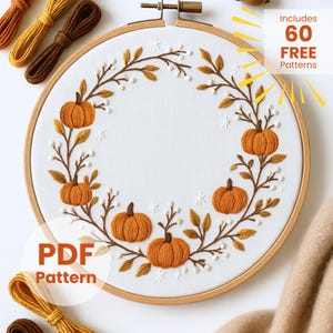 Pode incluir: Um bastidor de bordado com um fundo de tecido branco, decorado com um design outonal. O design apresenta abóboras laranja, galhos marrons e detalhes brancos. O texto "PDF Pattern" é visível. A imagem também inclui o texto "Includes 60 FREE Patterns".