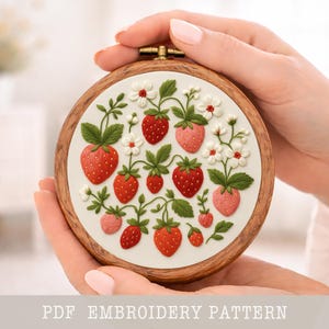 May include: A round embroidery hoop featuring a strawberry design. The design includes red and pink strawberries, green leaves, and white flowers on a white background. The hoop is held in hands and has a wooden frame. Text at the bottom reads "PDF EMBROIDERY PATTERN".