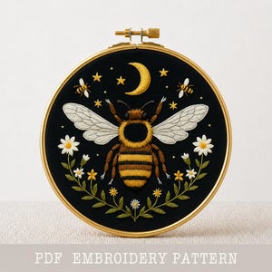 Puede incluir: Un bastidor de bordado redondo y negro con un diseño detallado de abeja. La abeja es amarilla y marrón con alas blancas, rodeada de estrellas, una luna creciente y detalles florales. El texto "PDF EMBROIDERY PATTERN" está en la parte inferior.