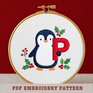 May include: Embroidered penguin holding a red letter "P" on white fabric, framed in a gold hoop. The design includes holly and berries. Text at the bottom reads "PDF EMBROIDERY PATTERN".