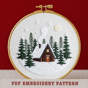 Op de afbeelding: Geborduurde winterscène in een gouden hoepel. Het ontwerp toont een met sneeuw bedekte hut met een rokende schoorsteen, omringd door groenblijvende bomen en sneeuwvlokken. De tekst "PDF EMBROIDERY PATTERN" staat onderaan.