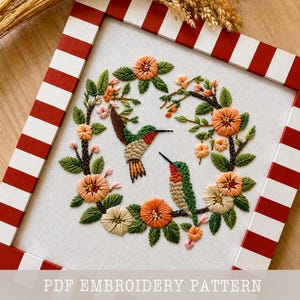 Könnte beinhalten: Gesticktes Kunstwerk mit zwei Kolibris, umgeben von einem floralen Kranz in Orange-, Grün- und Cremetönen. Das Kunstwerk ist in einem rot-weiß gestreiften Rahmen gerahmt. Der Text "PDF EMBROIDERY PATTERN" befindet sich unten.