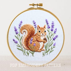 Puede incluir: Bordado de una ardilla sosteniendo una bellota, rodeada de ramitas de lavanda, en un aro dorado. La ardilla tiene pelaje marrón y una cola esponjosa. El texto "PDF EMBROIDERY PATTERN" está en la parte inferior.