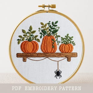 May include: A circular embroidery hoop containing white fabric with a hand-stitched design of three orange pumpkins on a brown wooden shelf. Green leaves sprout from the pumpkins, and a black spider hangs from a thread below the shelf. Text reads PDF EMBROIDERY PATTERN.