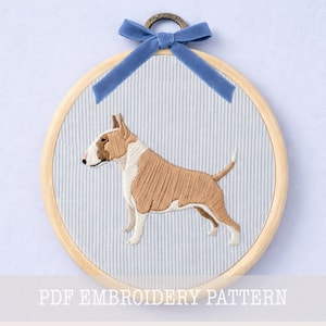 May include: A circular wooden embroidery hoop containing a fabric panel with a light blue and white pinstripe pattern. An embroidered tan and white bull terrier dog is centered on the fabric. A blue velvet ribbon is tied at the top metal hanger.