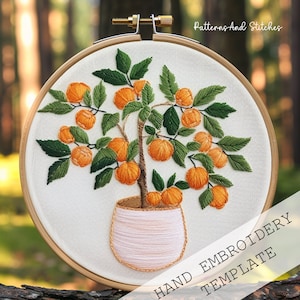 Op de afbeelding: Handborduurpatroon met een gepotte sinaasappelboom met groene bladeren en oranje vruchten. Het patroon wordt getoond in een houten borduurring.