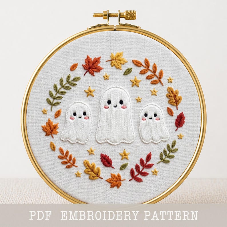 May include: A circular wooden embroidery hoop containing white fabric with a hand-stitched design of three white ghosts surrounded by a wreath of colorful autumn leaves and small yellow stars. The text PDF EMBROIDERY PATTERN is at the bottom.