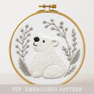 Pode incluir: Obra de arte bordada em um aro dourado com um urso polar branco com um olho e nariz pretos, cercado por detalhes florais cinza. O texto "PDF EMBROIDERY PATTERN" está na parte inferior. O bordado está em tecido branco.
